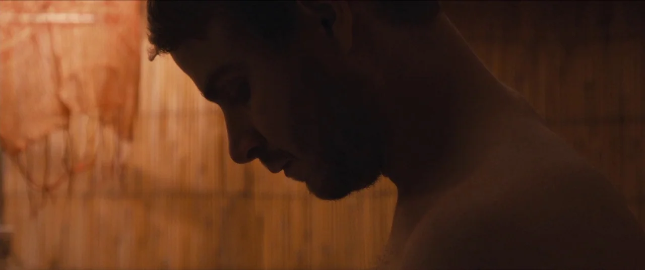 Simon Killer movie still: closeup — A dimly lit wooden structure with slats creating a textured background. A man is shirtless and captured in a…