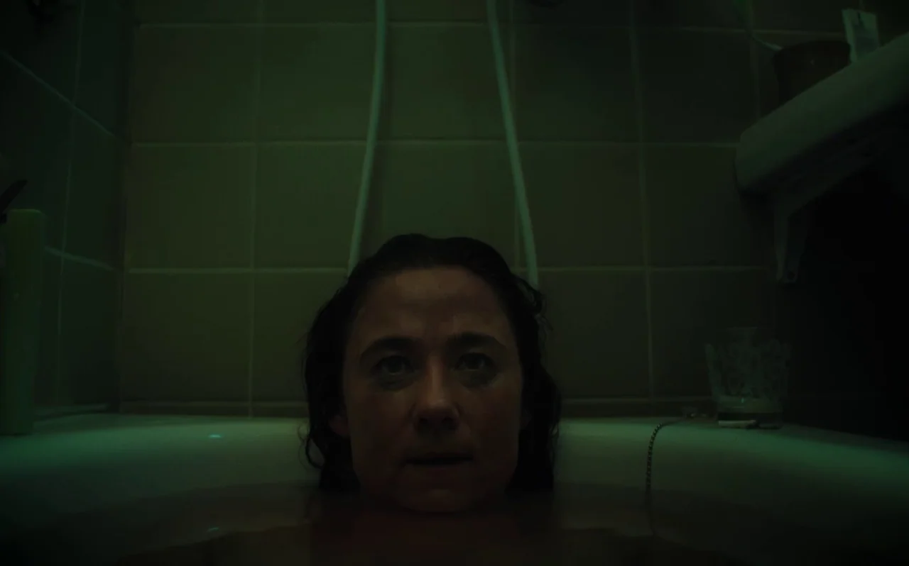 Knocking (2021) movie still: closeup — A dark, dimly lit bathroom features green-toned square tiles on the wall behind. A woman sits submerged in…