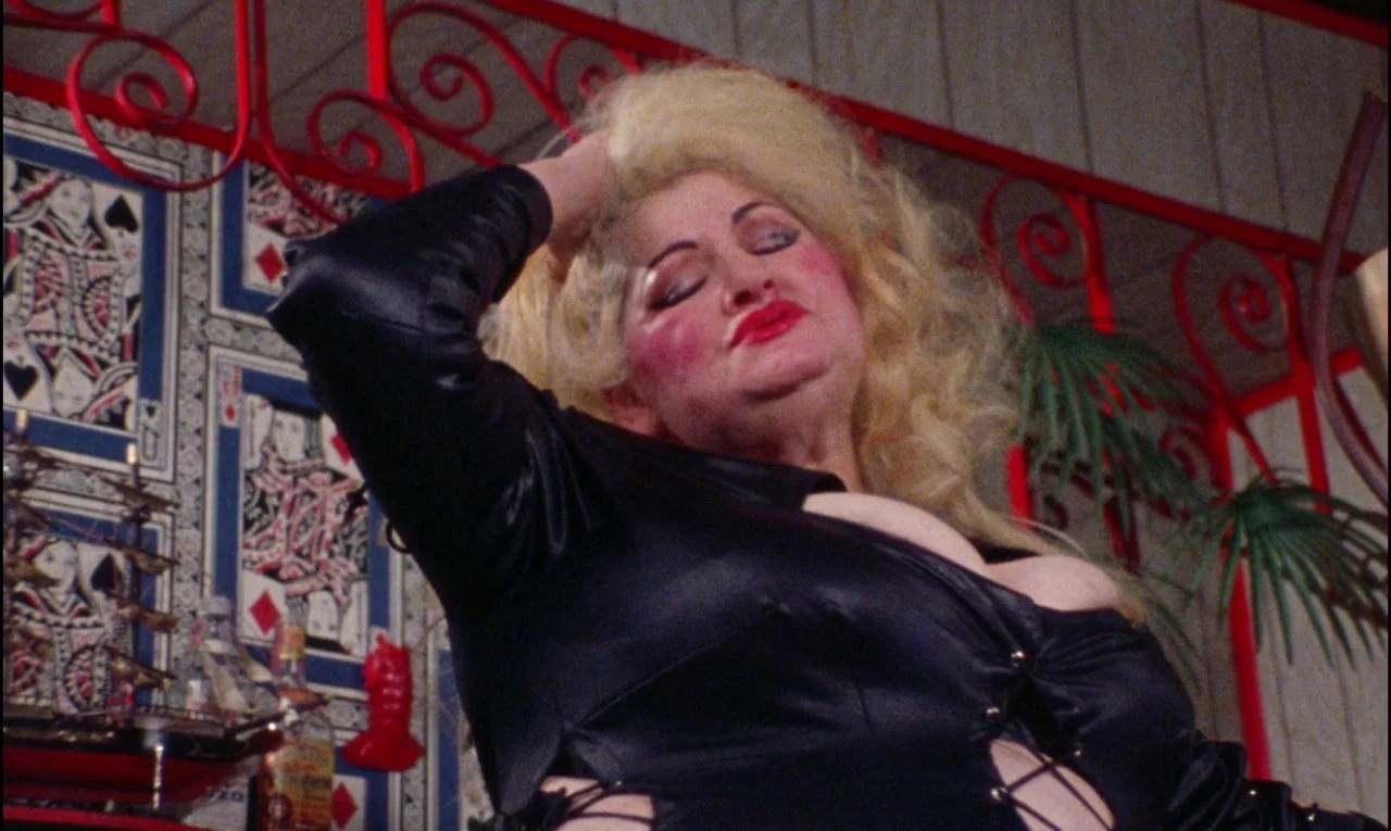 Female Trouble (1974) movie still: medium — A dimly lit room decorated with oversized playing card wall art and red scrollwork. A mature woman with…