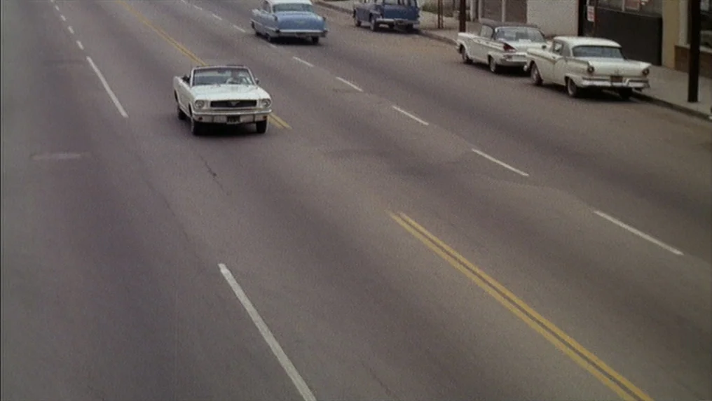 Targets (1968) movie still: wide — A paved asphalt road in an urban setting features multiple parked vintage automobiles. A white convertible moves…