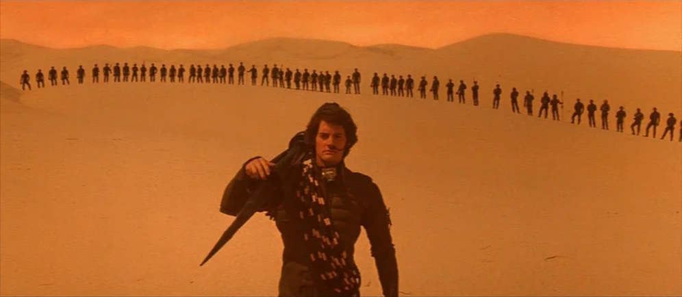 Dune (1984) movie still: fullbody — A vast sandy desert landscape stretches toward distant orange dunes. A man stands in the foreground while a long…
