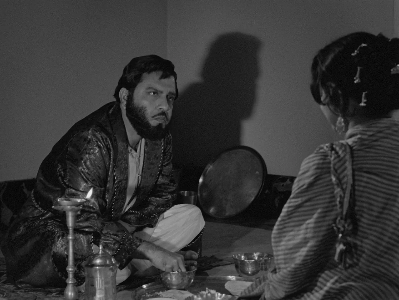 Charulata (1964) movie still: medium — A dimly lit room features a man and a woman sitting together on the floor. The man eats from a metal bowl by…