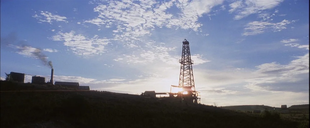 There Will Be Blood (2007) movie still: establishing — An oil derrick and processing buildings stand on a grassy hill during the day. Smoke rises…