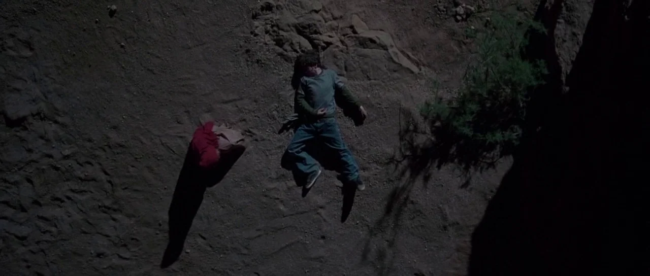 The Hills Have Eyes (2006) movie still: wide — A dry, rocky dirt surface at night with sparse desert vegetation. Two figures lie prone on the…