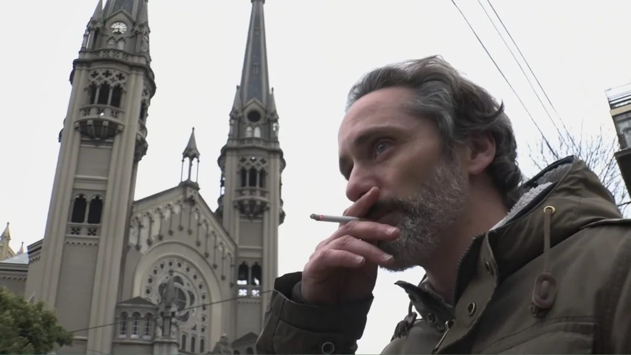 Smog in Your Heart (2022) movie still: medium — An ornate stone church with two high spires stands behind a man on the street. A man in a parka…