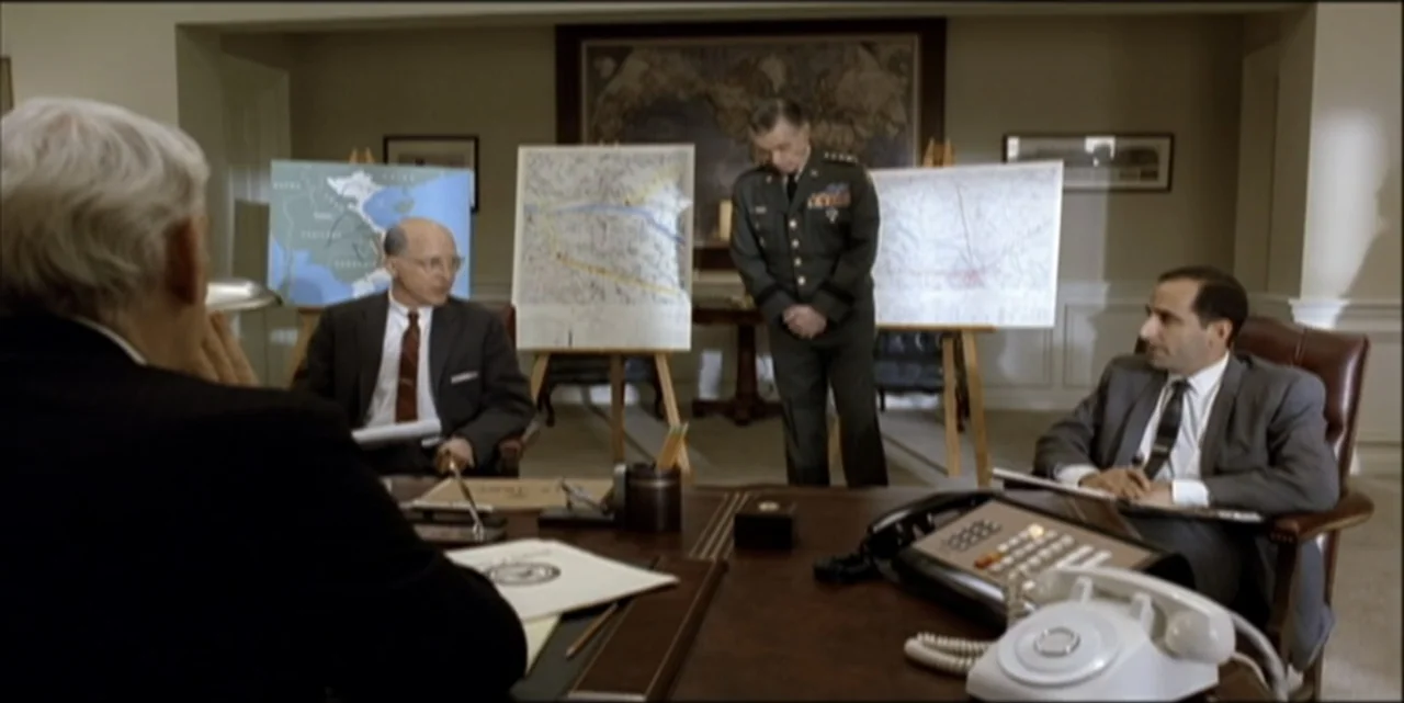 Path to War (2003) movie still: medium — A wood-paneled meeting room contains several easels displaying tactical maps for a presentation. Three men…