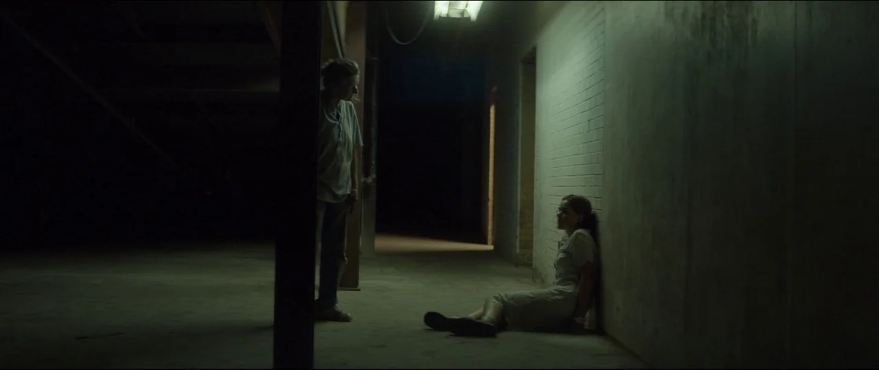 One & Two (2015) movie still: wide — A dimly lit industrial hallway features exposed brick walls and a concrete floor. A woman stands in the shadows…