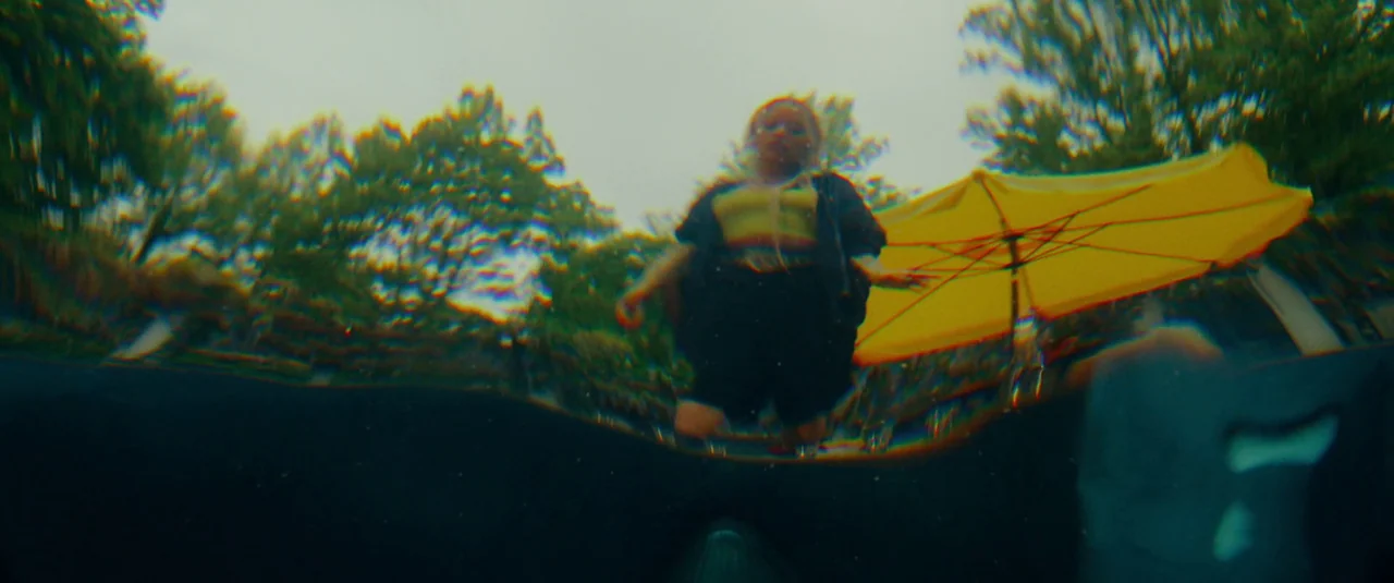 Bodies Bodies Bodies (2022) movie still: fullbody — A woman stands in water in a pool surrounded by tall green trees. The camera is submerged…