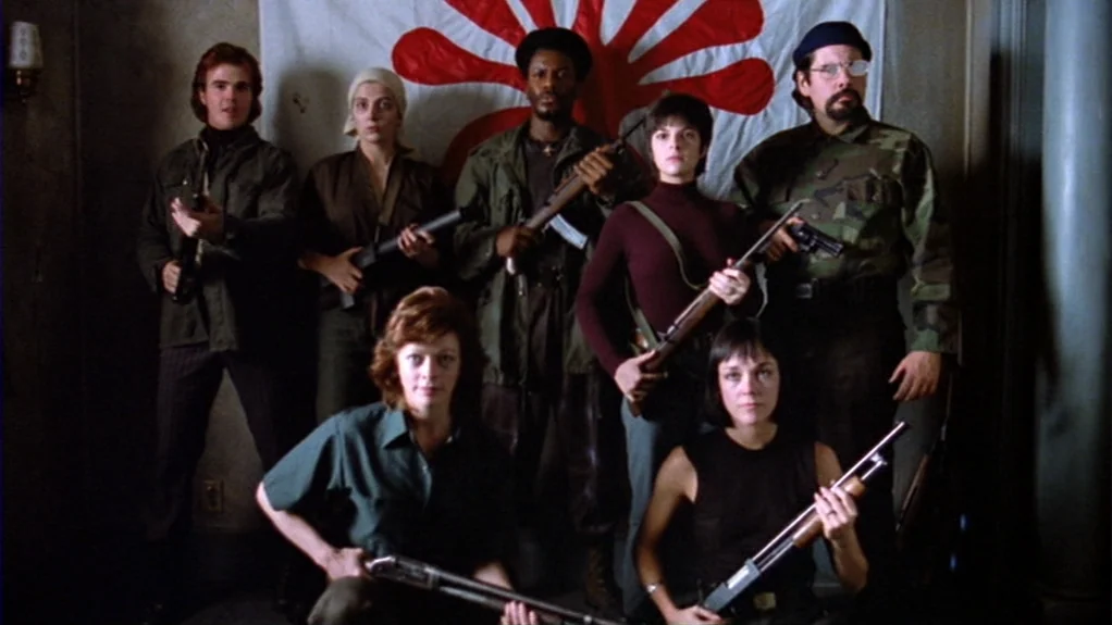 Patty Hearst (1988) movie still: fullbody — A group of seven people pose in front of a white flag with red rays. Each person carries a rifle or…