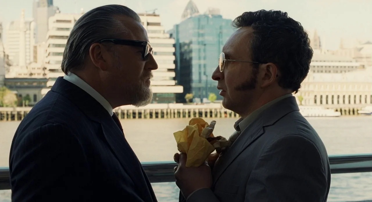 London Boulevard (2010) movie still: medium — Two men converse on a riverside pier with modern glass office buildings in background. One man holds a…