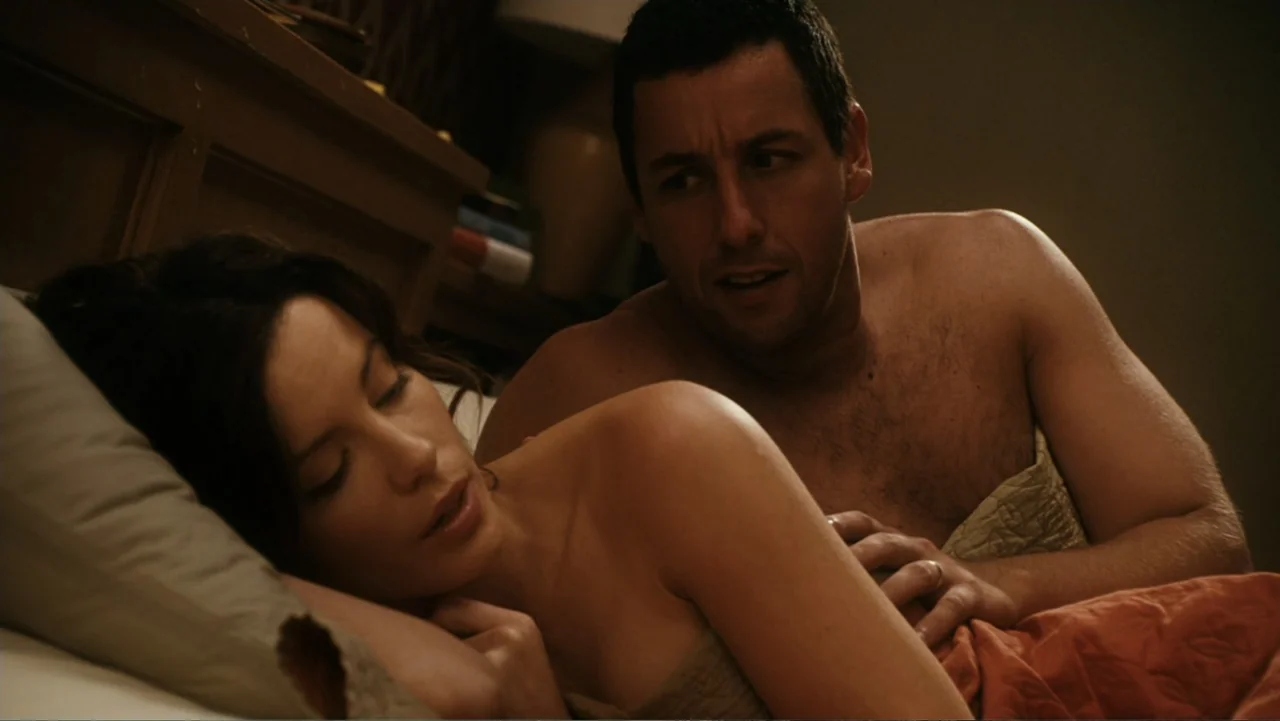 Click (2006) movie still: medium — A dimly lit bedroom features a wooden headboard and layered bed linens. A man and a woman rest together on a bed…