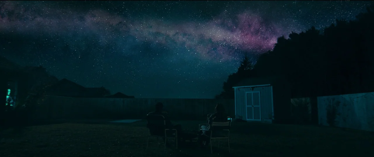 The Life of Chuck (2024) movie still: wide — A suburban residential backyard is visible at night under a vast, star-filled sky. Two figures sit in…