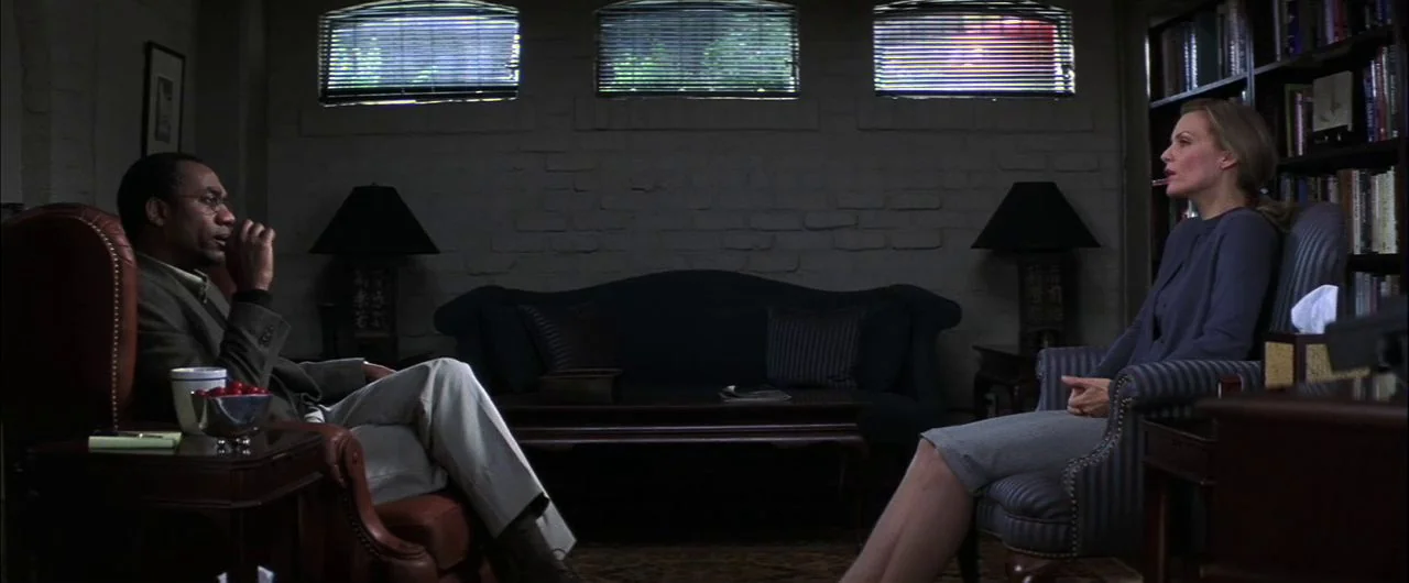 What Lies Beneath (2000) movie still: wide — A dimly lit room features a dark sofa flanked by matching table lamps. Two people sit in armchairs…