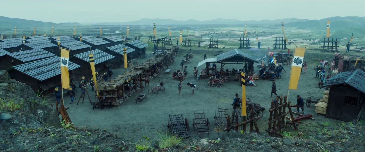 Kubi (2023) movie still: establishing — An expansive outdoor military camp features rows of wooden barracks and makeshift defensive structures. Many…