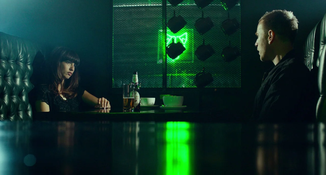 T2 Trainspotting (2017) movie still: medium — A dark booth with tufted leather benches faces a glowing green neon sign. A woman and man sit at the…