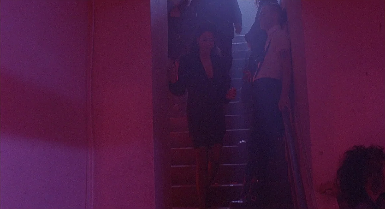 Less Than Zero (1987) movie still: medium — A dimly lit concrete stairwell is filled with dense haze and purple light. A woman walks down the steps…