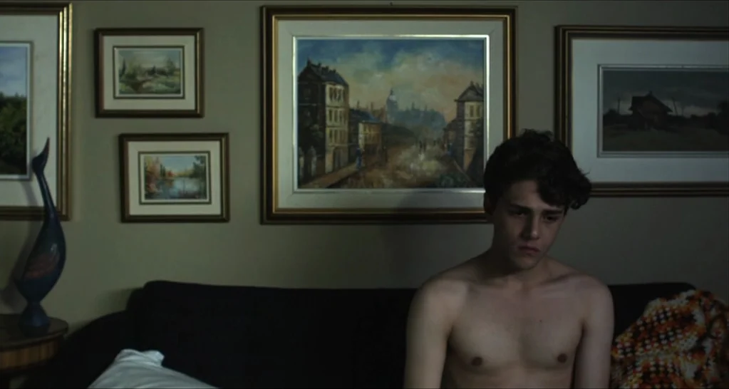 I Killed My Mother (J’ai tué ma mère) (2009) movie still: medium — A wall covered in various framed landscape oil paintings sits behind a dark…