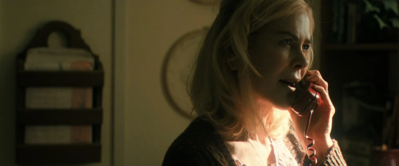Holland (2025) movie still: closeup — A dark, dimly lit interior room features a wooden wall-mounted letter holder. A middle-aged blonde woman holds…