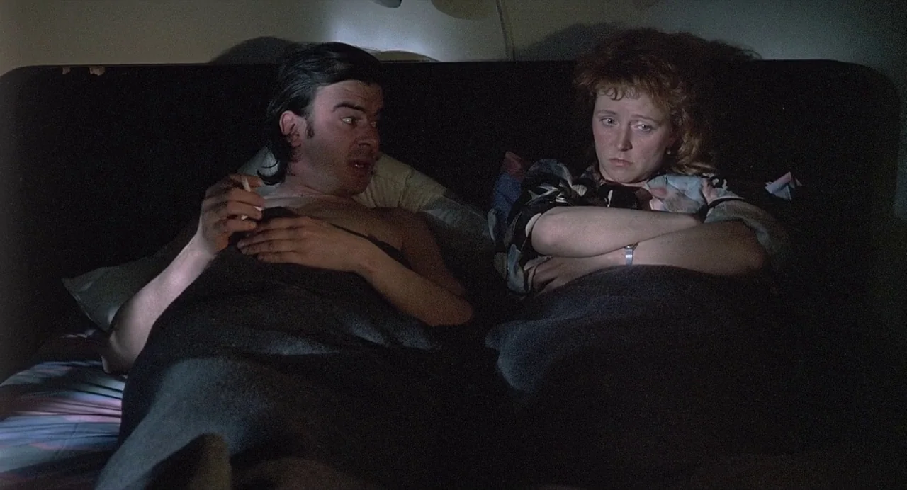 Ariel (1988) movie still: medium — A dimly lit room features a couple lying under blankets in a darkened space. The man gestures while speaking to…