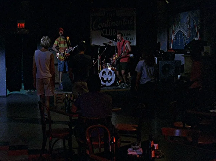 Slacker (1991) movie still: wide — A dimly lit club interior with a small stage and scattered dark wooden tables. A two-piece band plays on the…