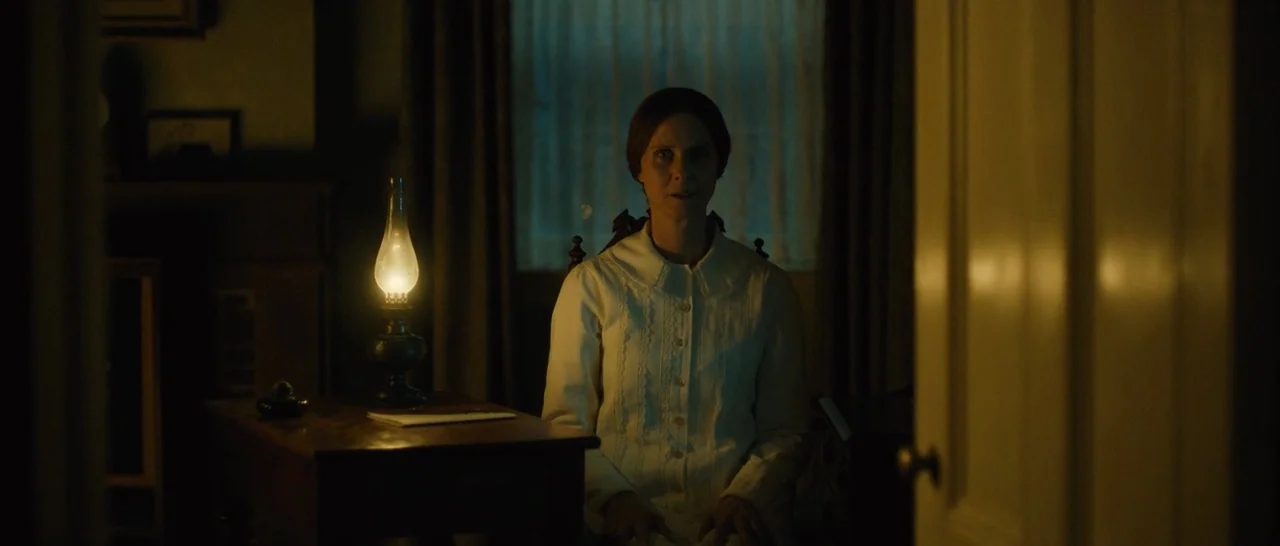 A Quiet Passion (2016) movie still: medium — A dimly lit room features a woman seated by a glowing oil lamp. She remains still while looking forward…