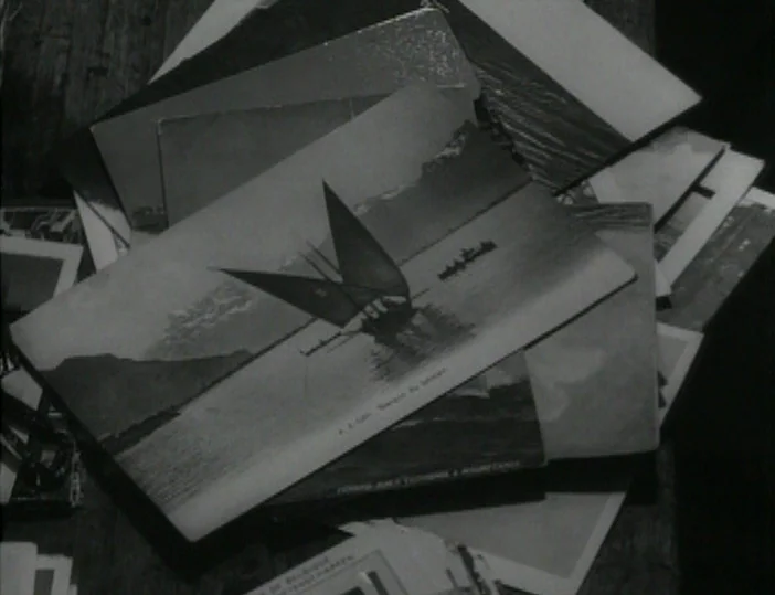Les Carabiniers (1963) movie still: closeup — A pile of various vintage postcards lies scattered across a wooden tabletop surface. A prominent…