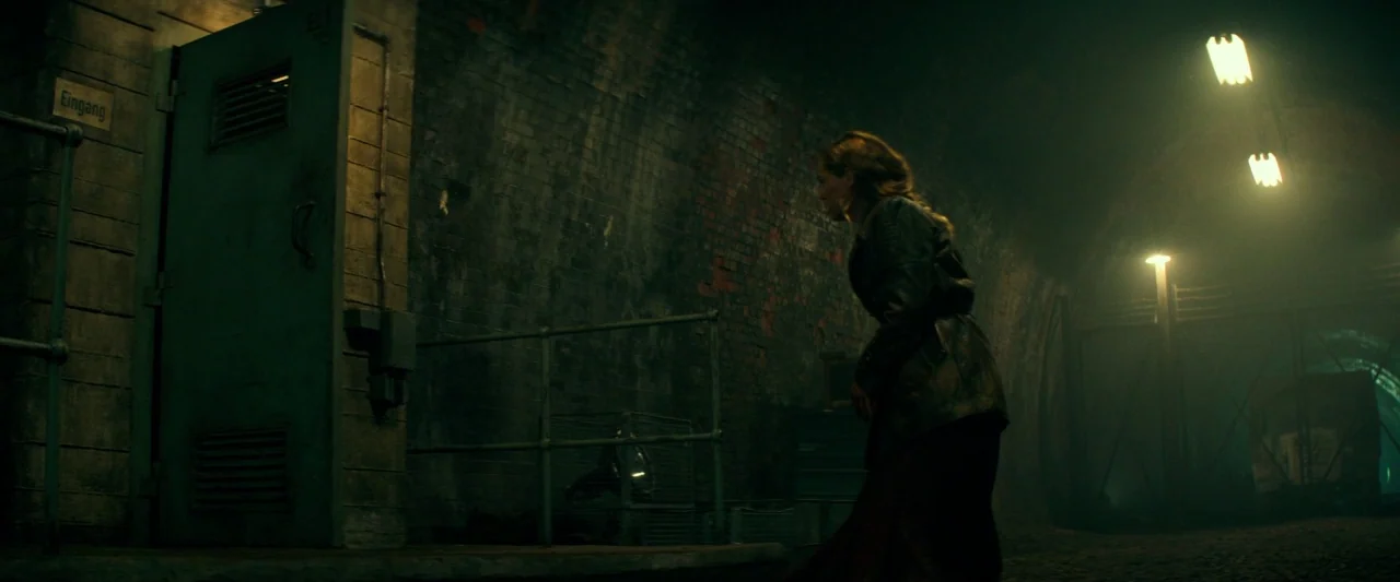 Overlord (2018) movie still: medium — A dark underground brick tunnel features a heavy metal door labeled with German signage. A woman moves through…