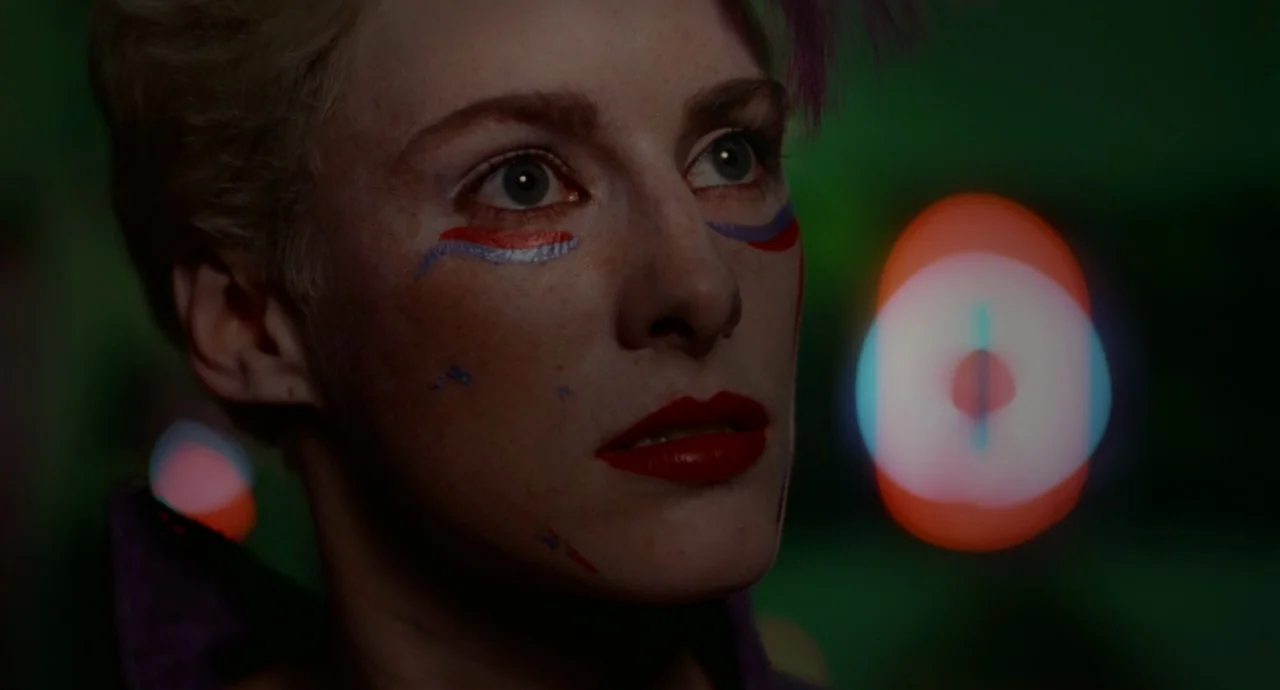 Liquid Sky (1982) movie still: closeup — A dimly lit space features a woman with elaborate face paint against a dark background. Red and blue bokeh…