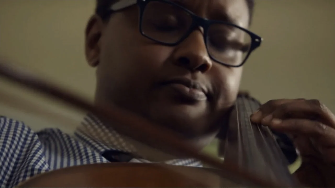 Suited (2016) movie still: closeup — A dimly lit indoor space with neutral-colored walls in the background. A man plays the cello with intense focus…