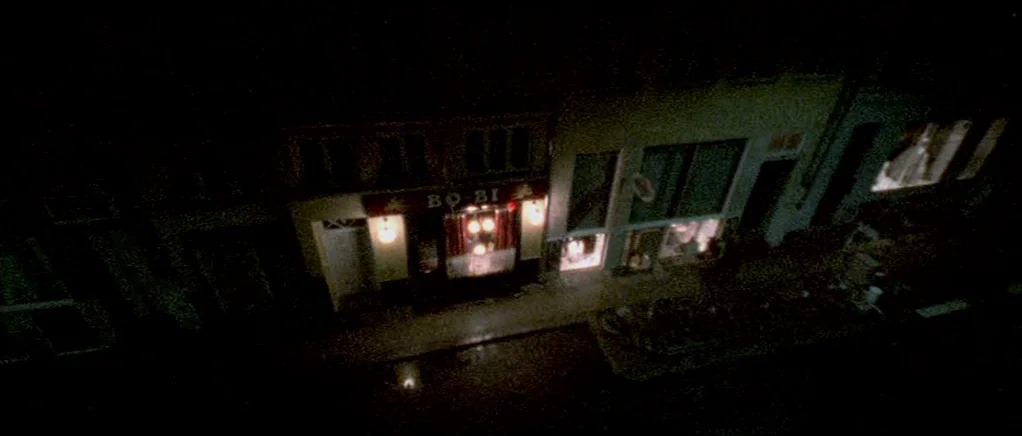 Reconstruction (2003) movie still: establishing — A nighttime view looking down at a row of illuminated storefronts on a street. Glass windows…