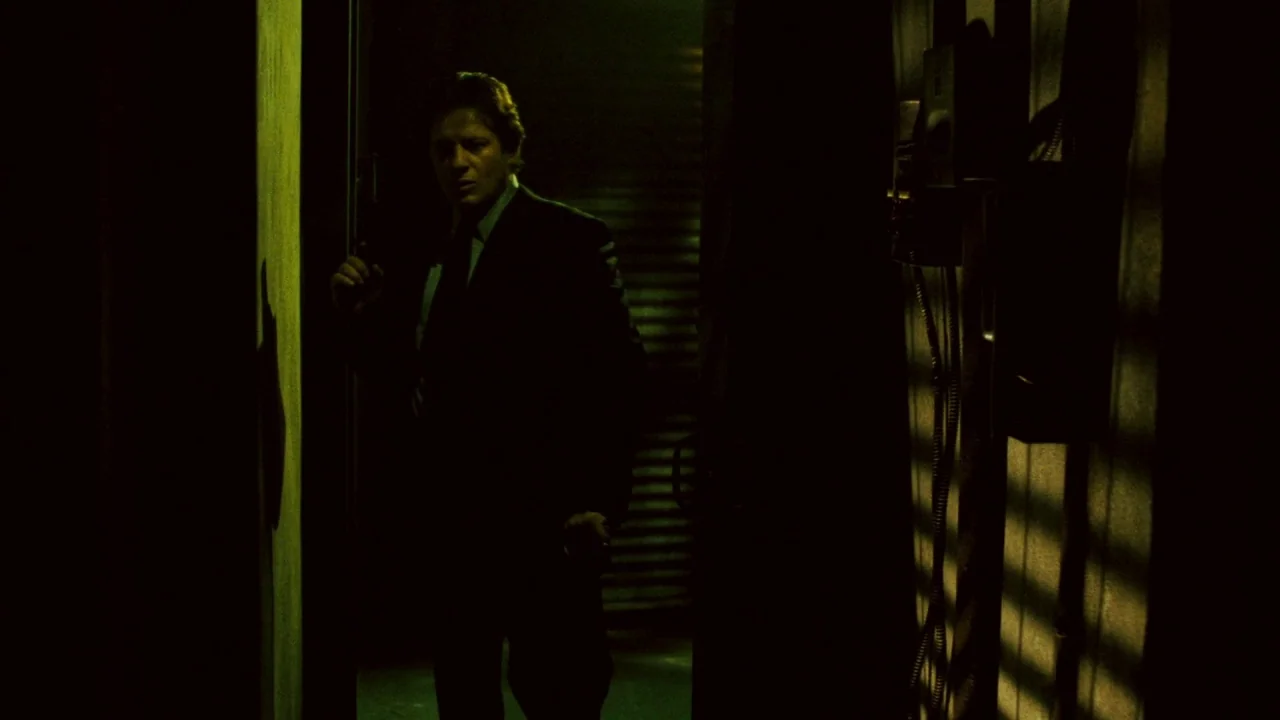 Saw V (2008) movie still: medium — A dark, narrow corridor is illuminated by green-tinted light filtering through horizontal window blinds. A man in…