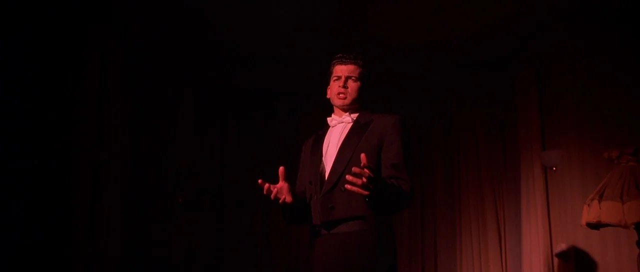 Heavenly Creatures (1994) movie still: medium — A man in formal wear stands on a dimly lit stage during a performance. He gestures with his open…
