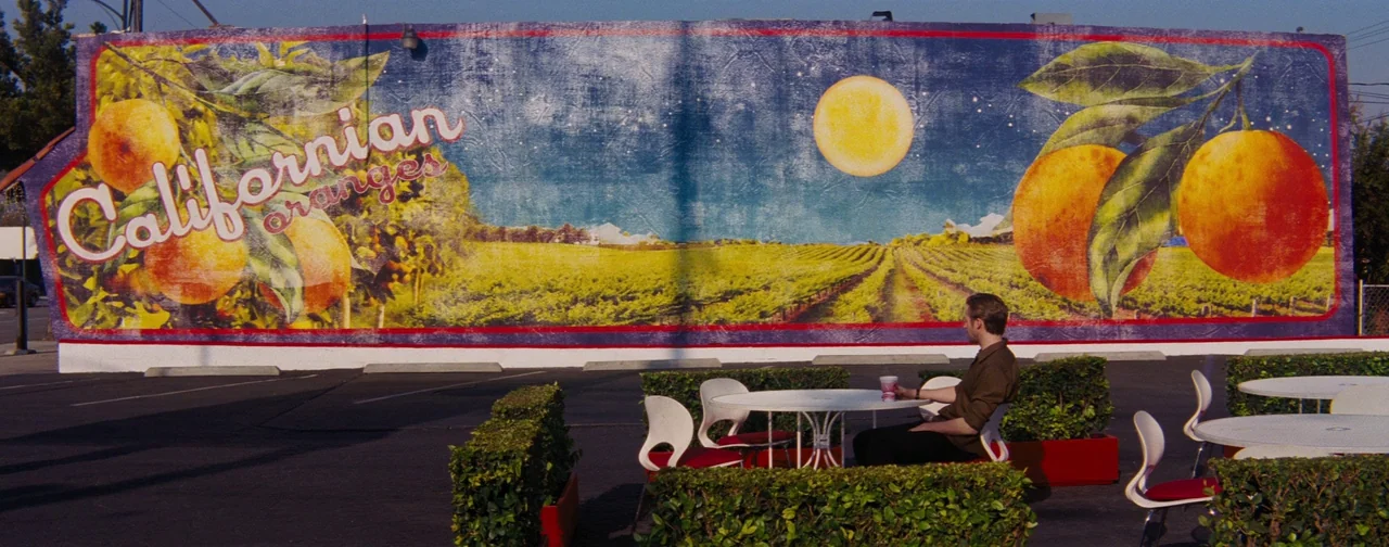 La La Land (2016) movie still: wide — A sprawling parking lot features a large, colorful mural painted on a long wall. A man sits alone at an…