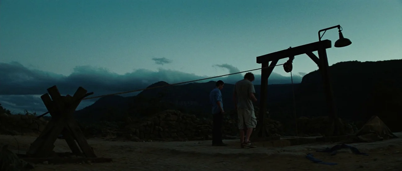 The Ruins (2008) movie still: wide — An open landscape at dusk features a wooden well structure under a cloudy sky. Two men stand beside the well,…