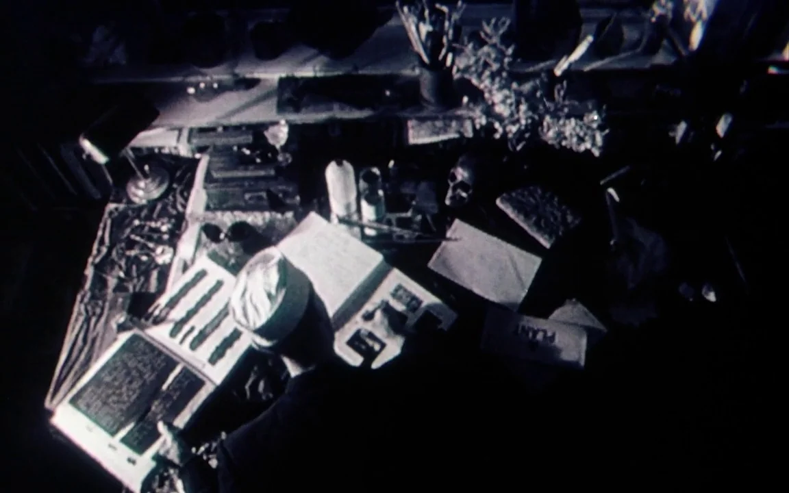 The Last of England (1987) movie still: medium — A dark study cluttered with various tools and a human skull. A person sits at the desk reading and…