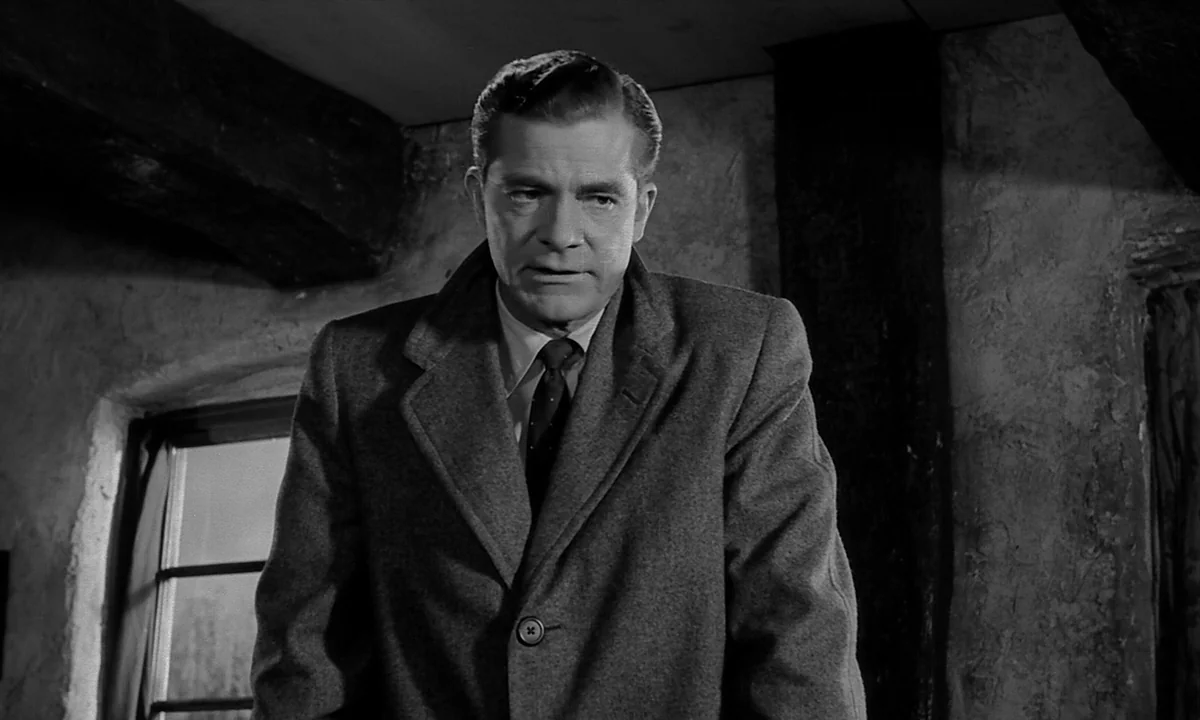 Night Of The Demon (1957) movie still: medium — A rustic interior features stone walls, wooden ceiling beams, and a small window. A middle-aged man…