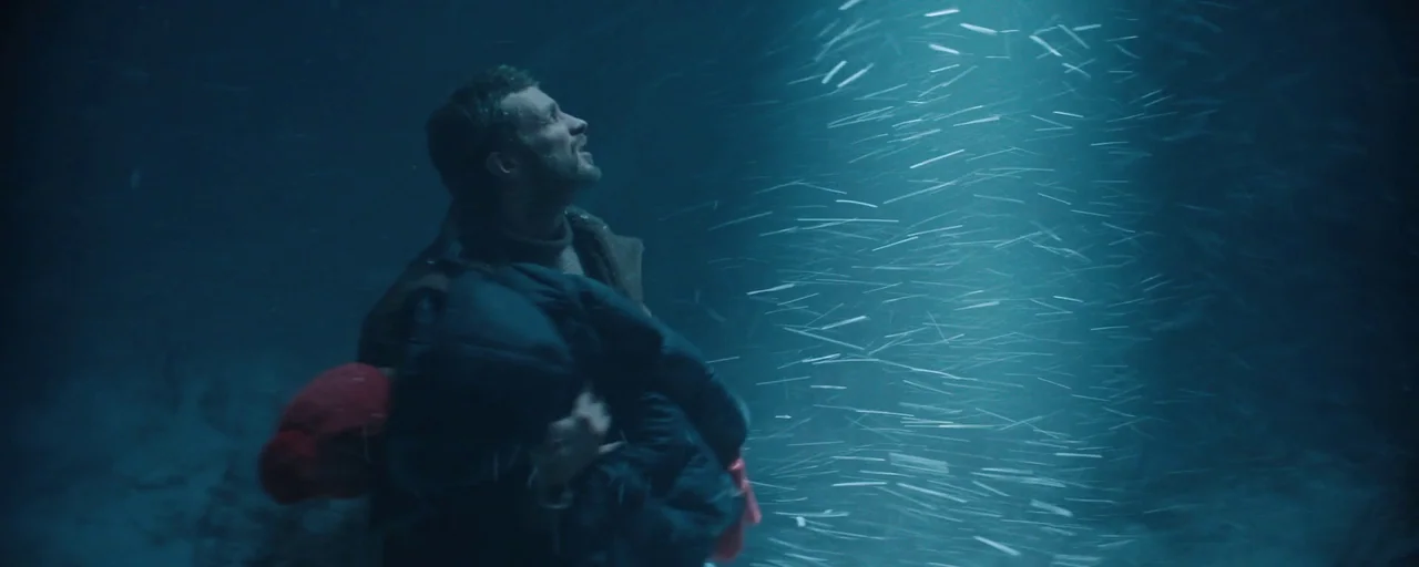 Petrov’s Flu (2021) movie still: medium — A dark, snow-filled night scene outdoors with limited visibility. A man carrying a bundled figure walks…