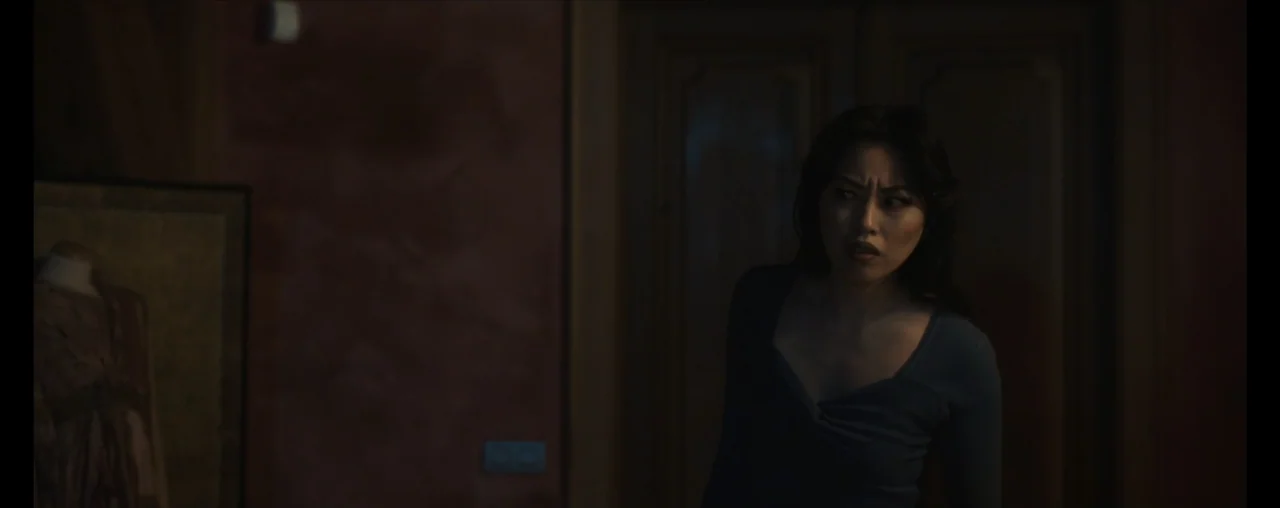 The Wrong Tenant (2022) movie still: medium — A dimly lit interior space with dark walls and a recessed doorway. A young woman with dark hair stands…
