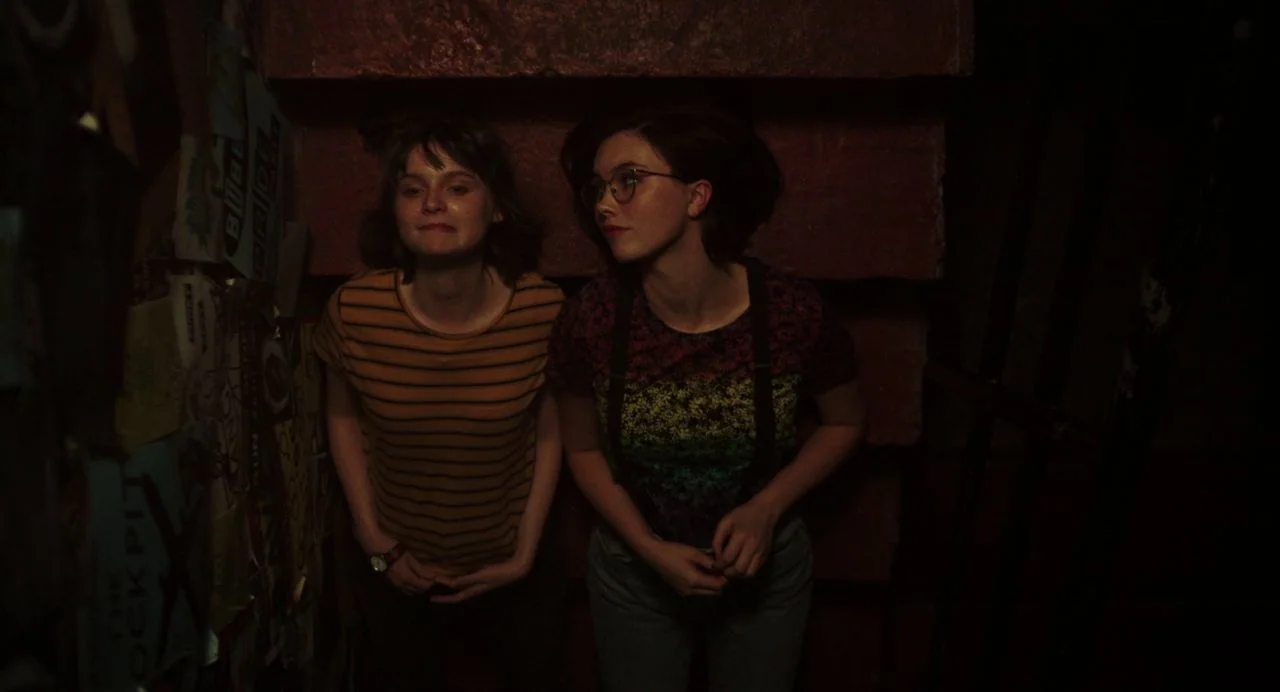 Dating Amber movie still: medium — A dimly lit stairwell features a dark red wall covered in layers of posters. Two young women stand close together…