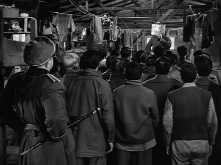 Stalag 17 (1953) movie still: wide — A crowded wooden structure with clothing hanging from wires stretched across the rafters. A large group of men…