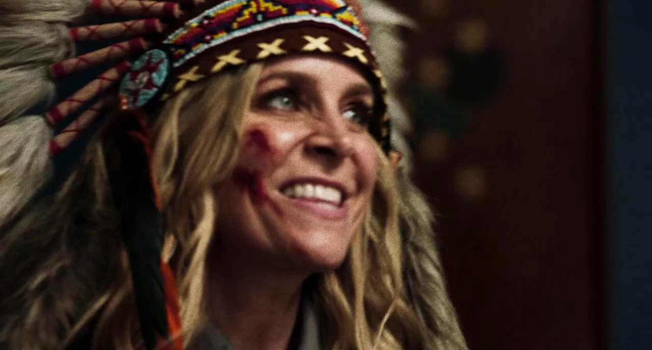 3 From Hell (2019) movie still: closeup — A woman wears a traditional feathered headdress in a dimly lit room. She has red paint streaks on her…
