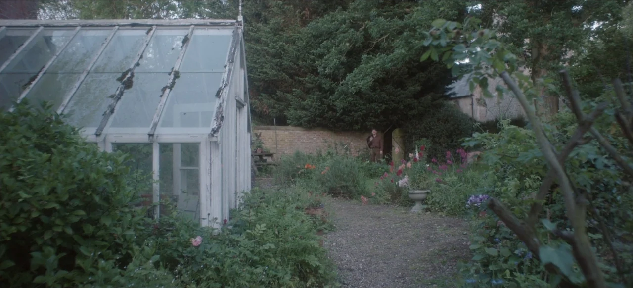We Go In at Dawn (2020) movie still: wide — A dense garden with a white greenhouse structure and overgrown evergreen trees. A lone man stands on a…