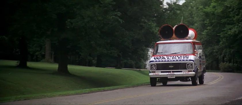 Nashville (1975) movie still: wide — A paved road winds through a lush, dense forest with manicured green grassy hillsides. A vintage Chevrolet…
