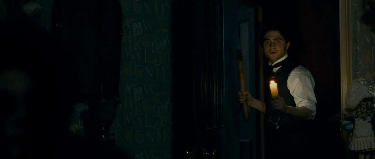 The Woman in Black (2012) movie still: medium — An old hallway features dark blue wallpaper covered in faded alphabet prints. A man holds a lit…