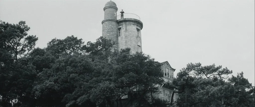 Les Créatures (1966) movie still: wide — A stone lighthouse rises above a dense cluster of mature trees under grey skies. A lone person stands on…