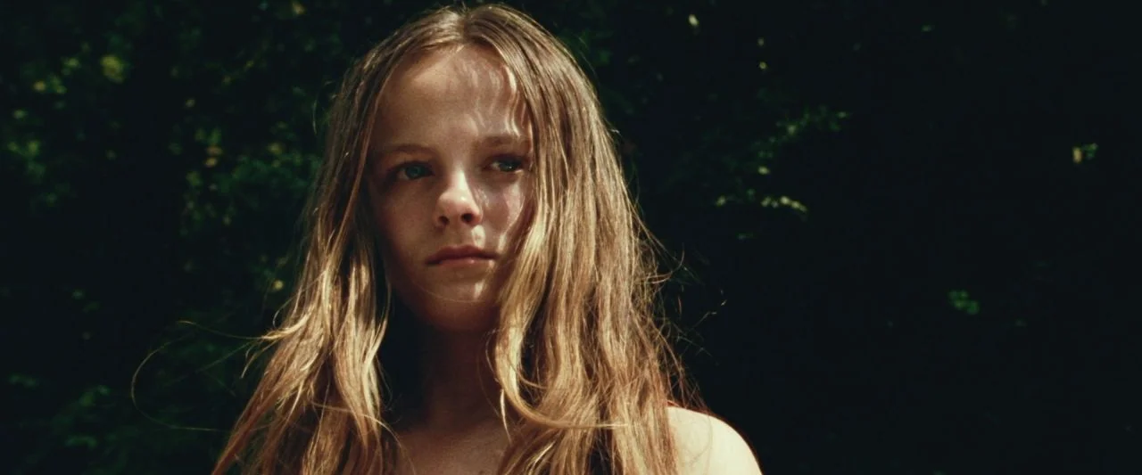 Adoration (2019) movie still: closeup — A teenage girl stands outdoors surrounded by dense, dark green foliage under trees. Sunlight illuminates her…
