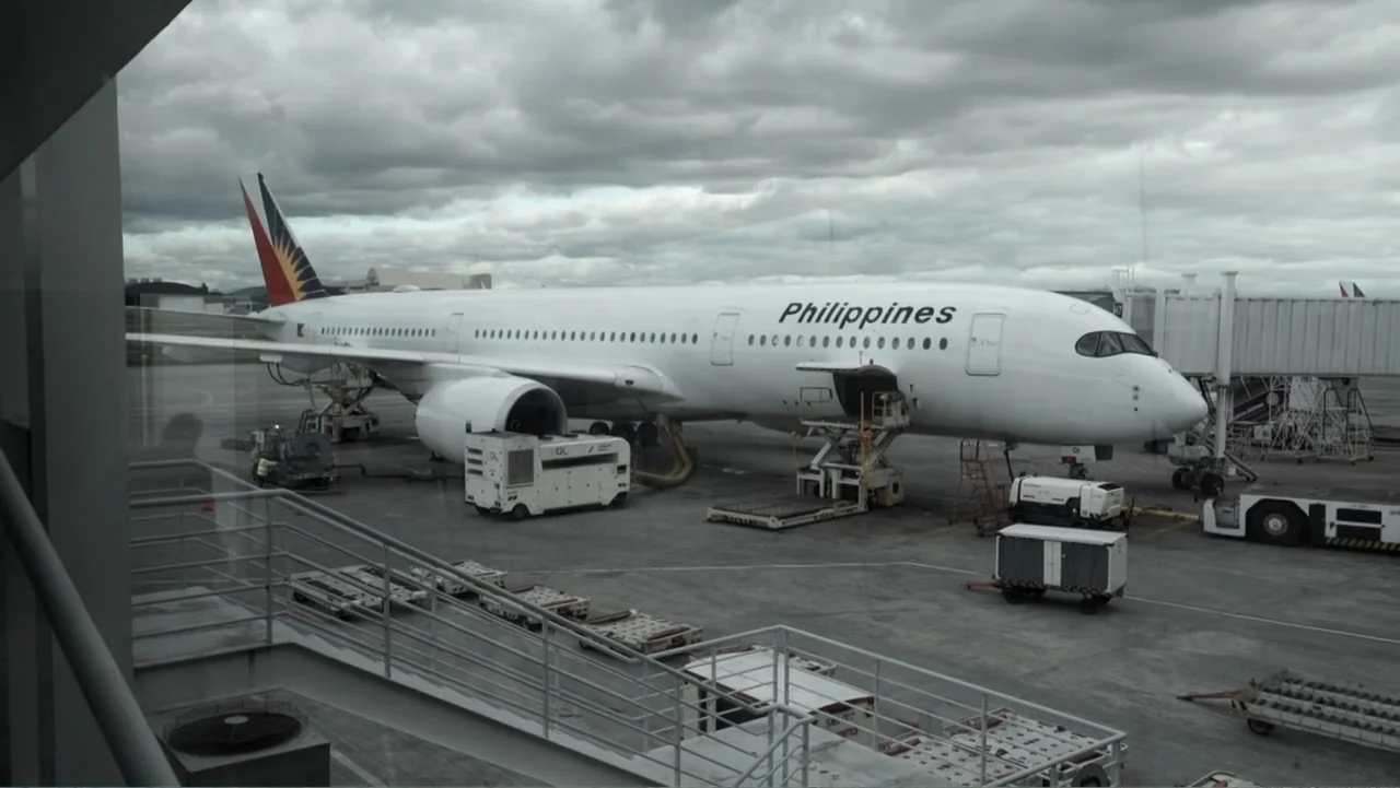 Escape From Saudi (2019) movie still: wide — A white passenger jet rests on a concrete tarmac under heavy overcast skies. Ground service equipment…