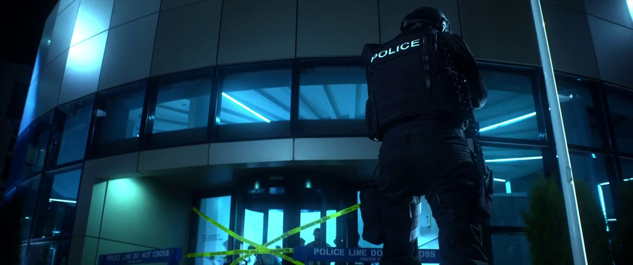 Mayhem (2017) movie still: medium — A curved modern glass office building is cordoned off with yellow quarantine police tape. A lone tactical police…