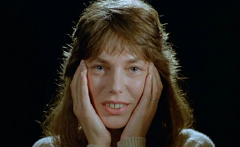 Jane B. par Agnès V. (1988) movie still: closeup — A woman stands against a black background in an studio setting. She presses her hands against…