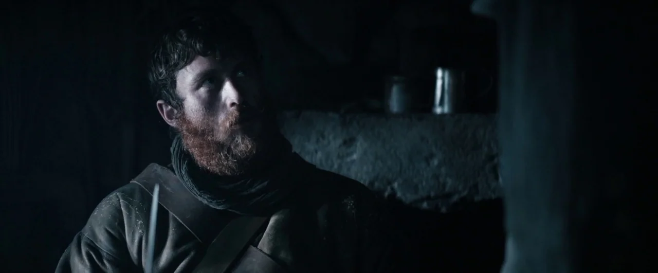 Black ’47 (2018) movie still: medium — A dark, sparsely lit interior space with rough masonry walls and metallic items visible. A bearded man with a…